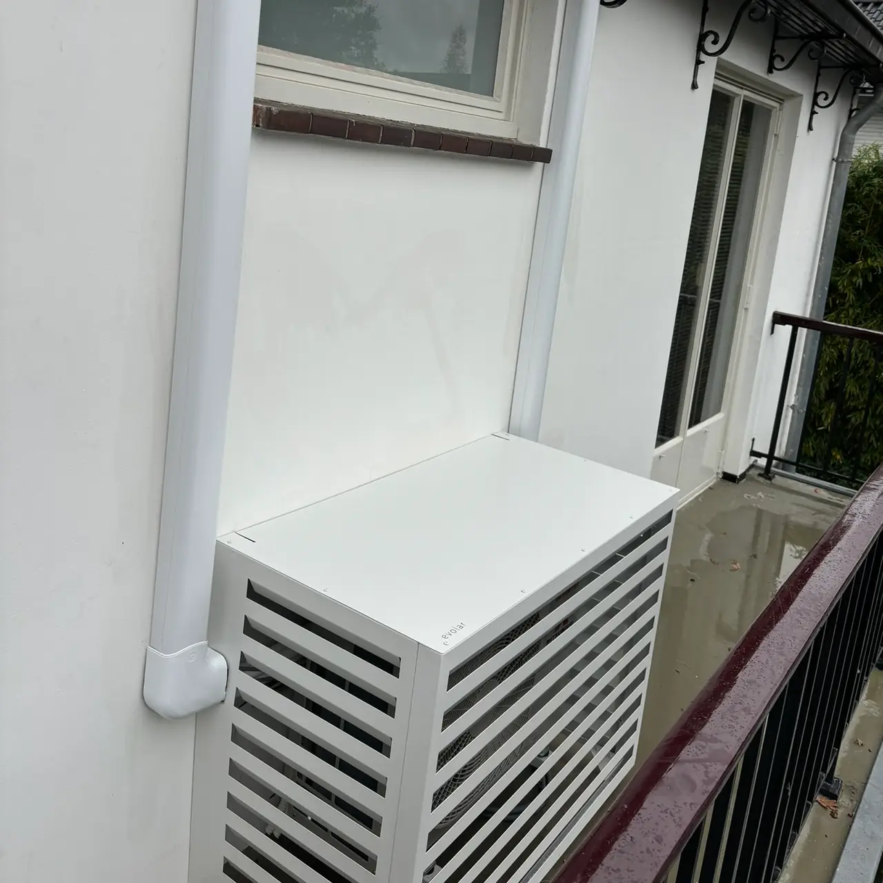 An outdoor unit in a white louvred cover on a balcony, with the pipework in a white duct along the wall.