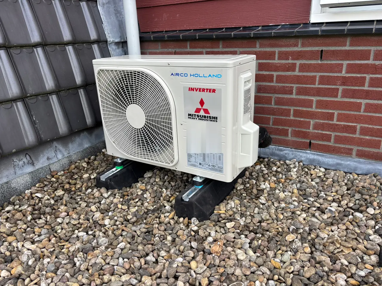 Een buitenunit van Mitsubishi Heavy Industries op het grind van een dak, tussen een pannendak en een bakstenen gevel.