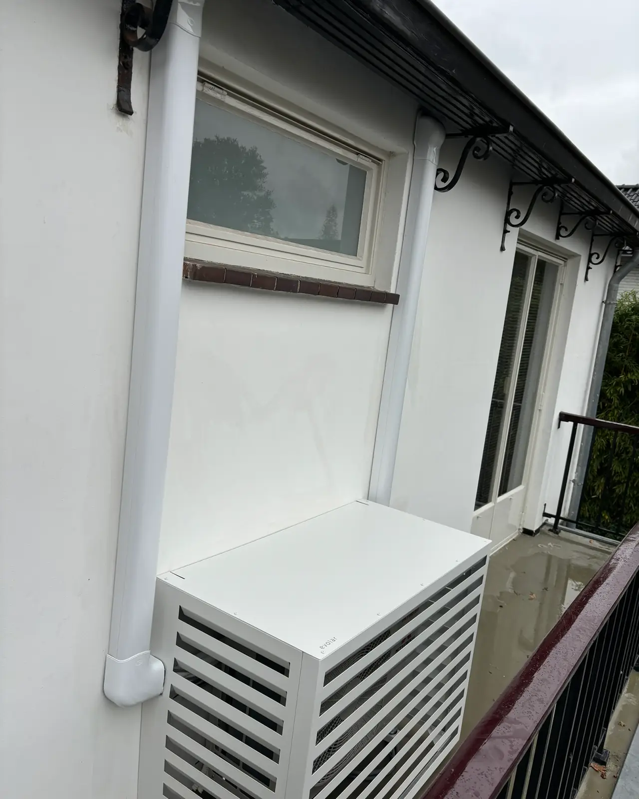 Een buitenunit in een witte lamellenkast op een balkon, met de leiding in een witte goot langs de gevel.