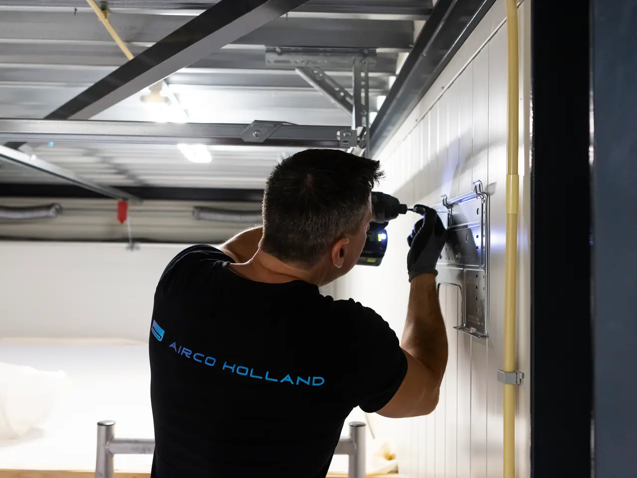 Peter, seen from behind with Airco Holland on his shirt, drills the mounting plate for the indoor unit in place.