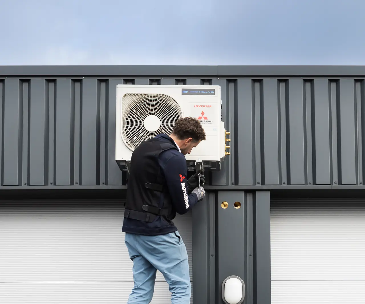 Rein staat op een rolsteiger en werkt aan een nieuwe buitenunit aan de gevel van een bedrijfspand.