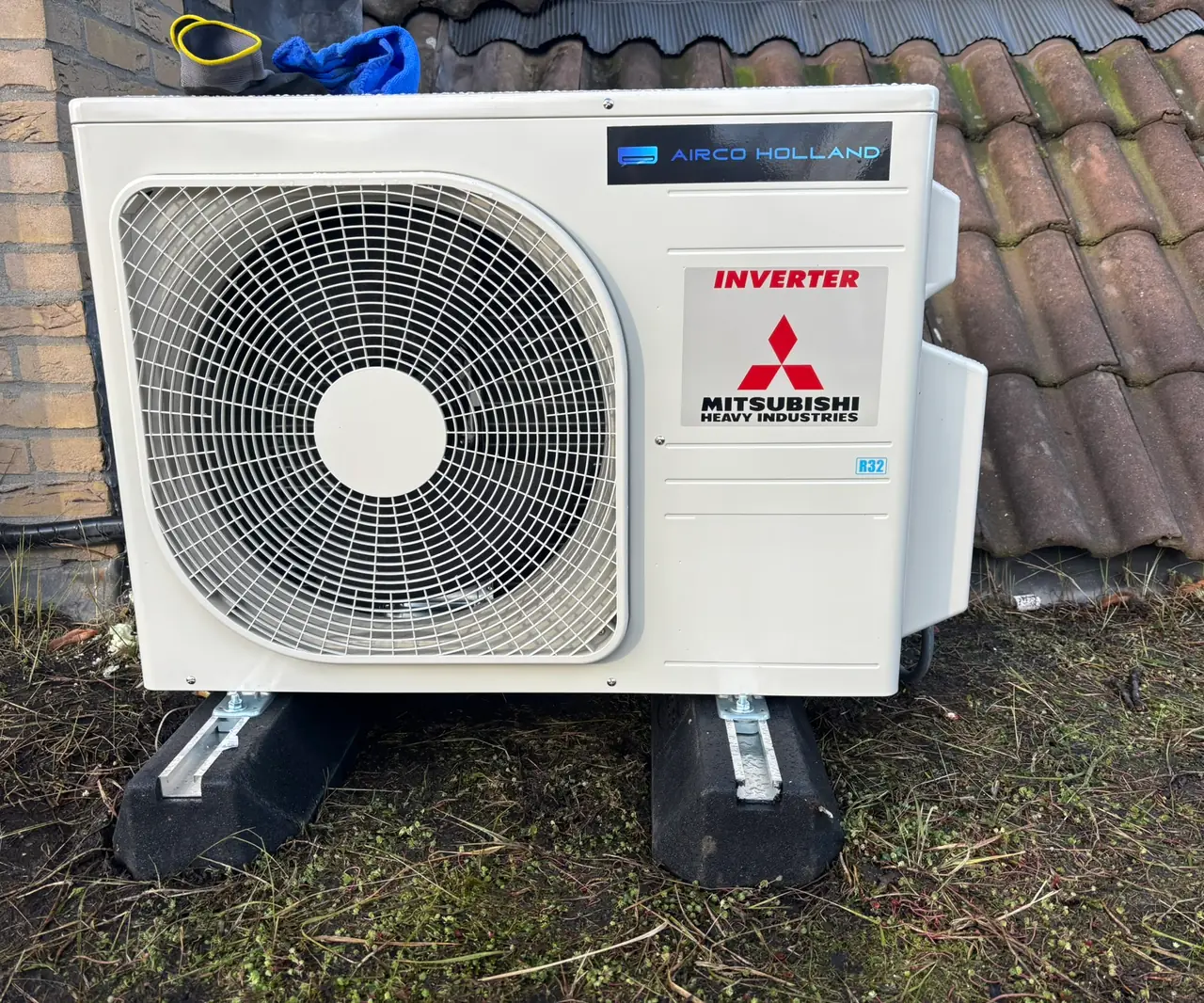 Een buitenunit op rubberen voeten naast een pannendak en baksteen.