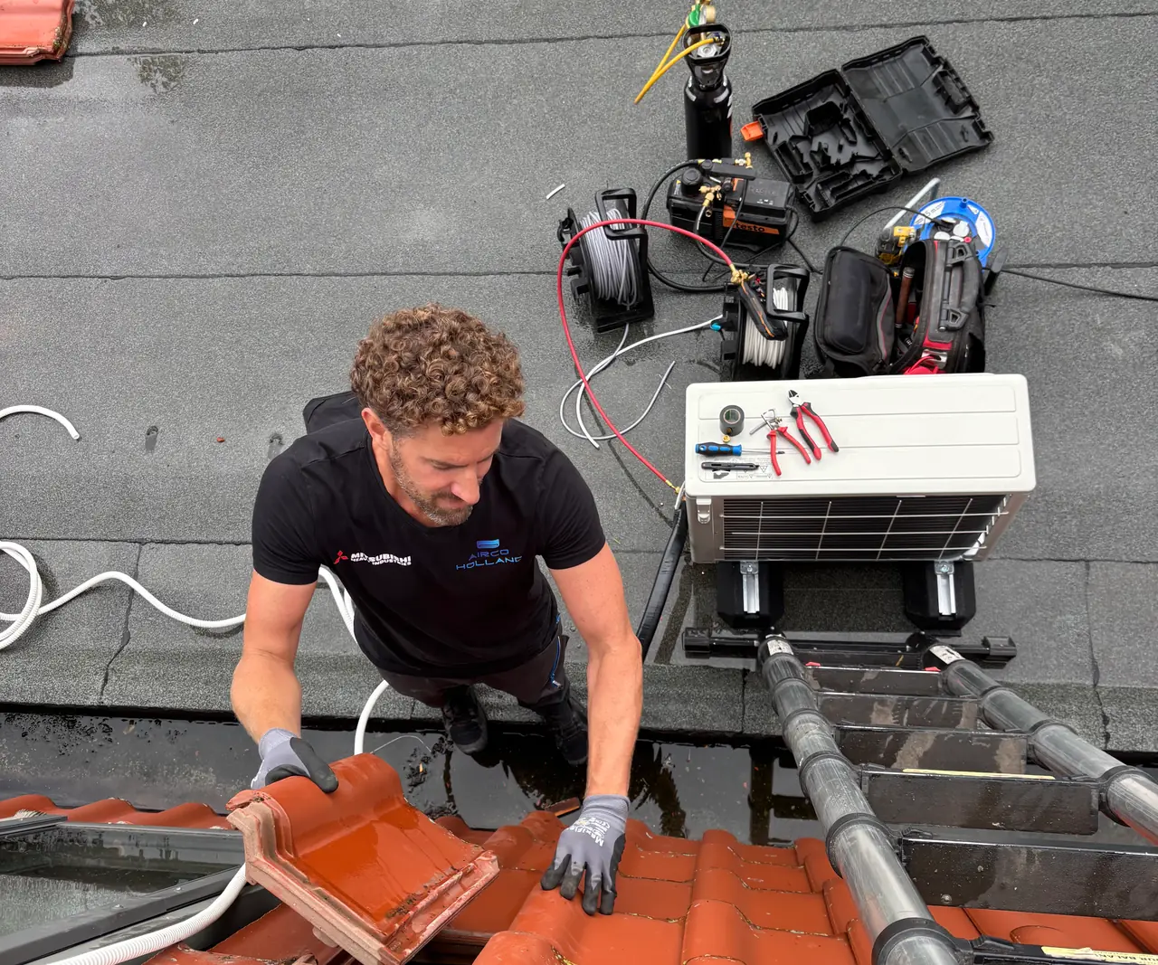 Van bovenaf: Rein legt de dakpannen terug over de leiding van de airco, met de buitenunit en zijn gereedschap erachter.