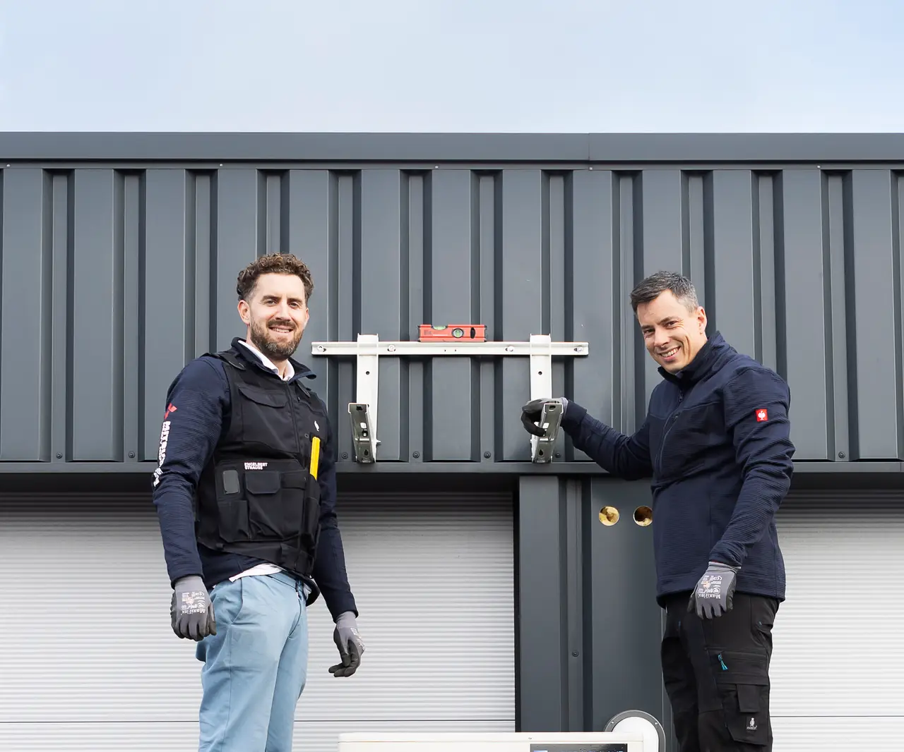 Rein en Peter staan op de rolsteiger bij de nieuwe buitenunit en lachen in de camera.