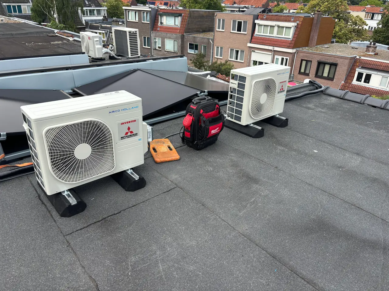 Twee nieuwe buitenunits met een gereedschapstas op een plat dak, met de achtergevels van een rij huizen.