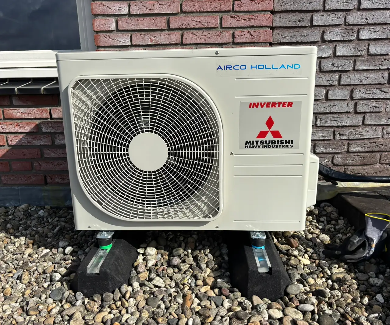 Een buitenunit met de stickers van Airco Holland en Mitsubishi Heavy Industries op dakvoeten in grind.