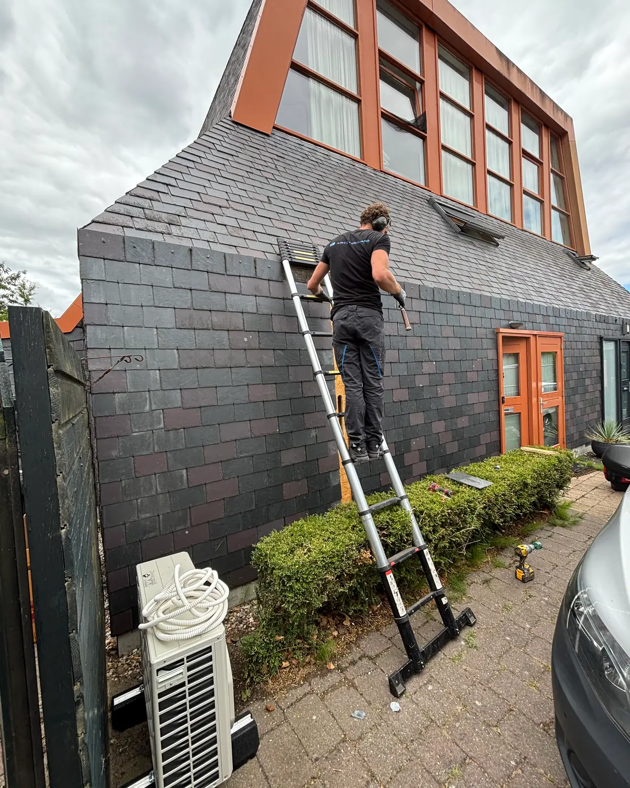 Rein staat op een ladder tegen de leien gevel van een opvallende woning met oranje kozijnen.