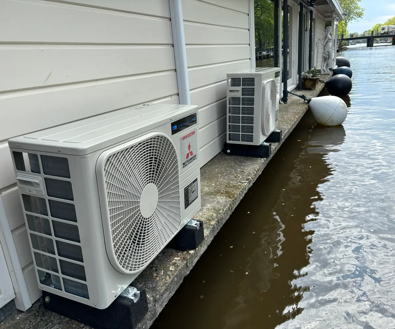 Twee buitenunits op de rand van een woonboot aan een gracht, met een brug op de achtergrond.