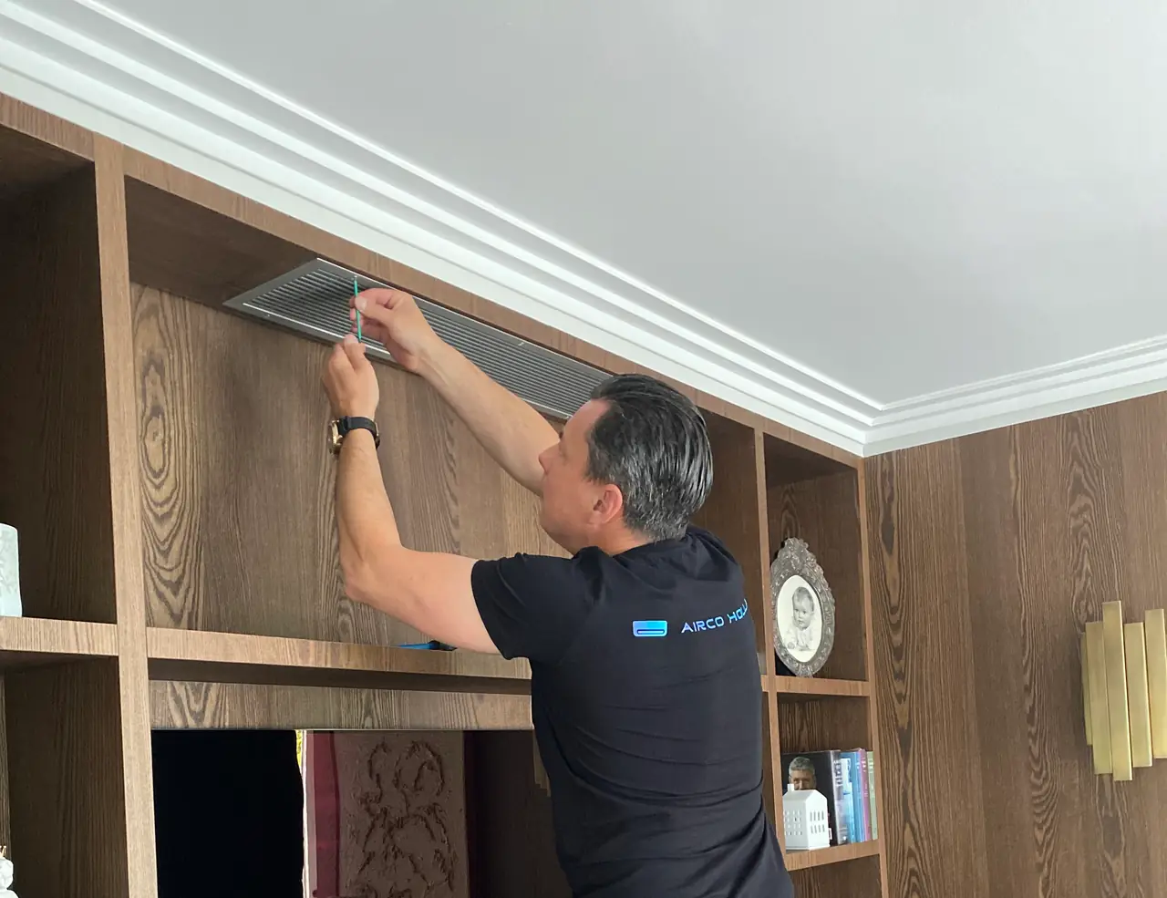 Peter stelt op een trapje het rooster af van een airco die boven in een houten maatwerkkast zit.