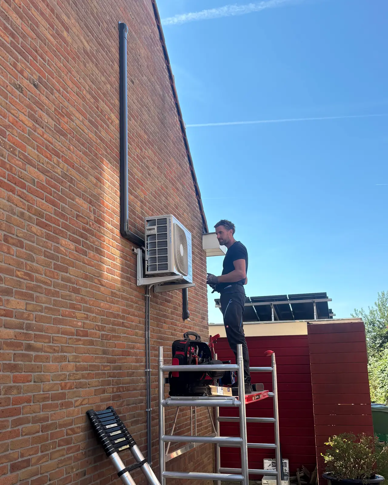 Rein op een rolsteiger bij een nieuwe buitenunit hoog aan een bakstenen kopgevel, met een zwarte leidinggoot.