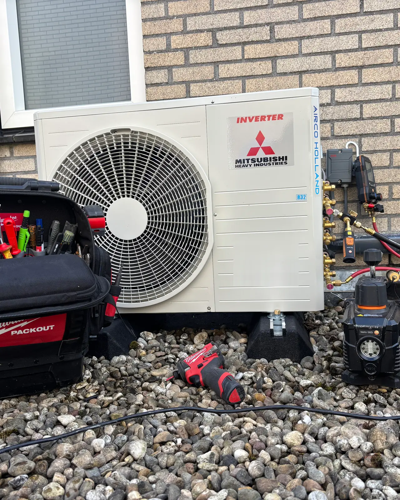 Een nieuwe buitenunit op een grinddak met een gereedschapstas, een vacuümpomp en een meter aan de kranen.