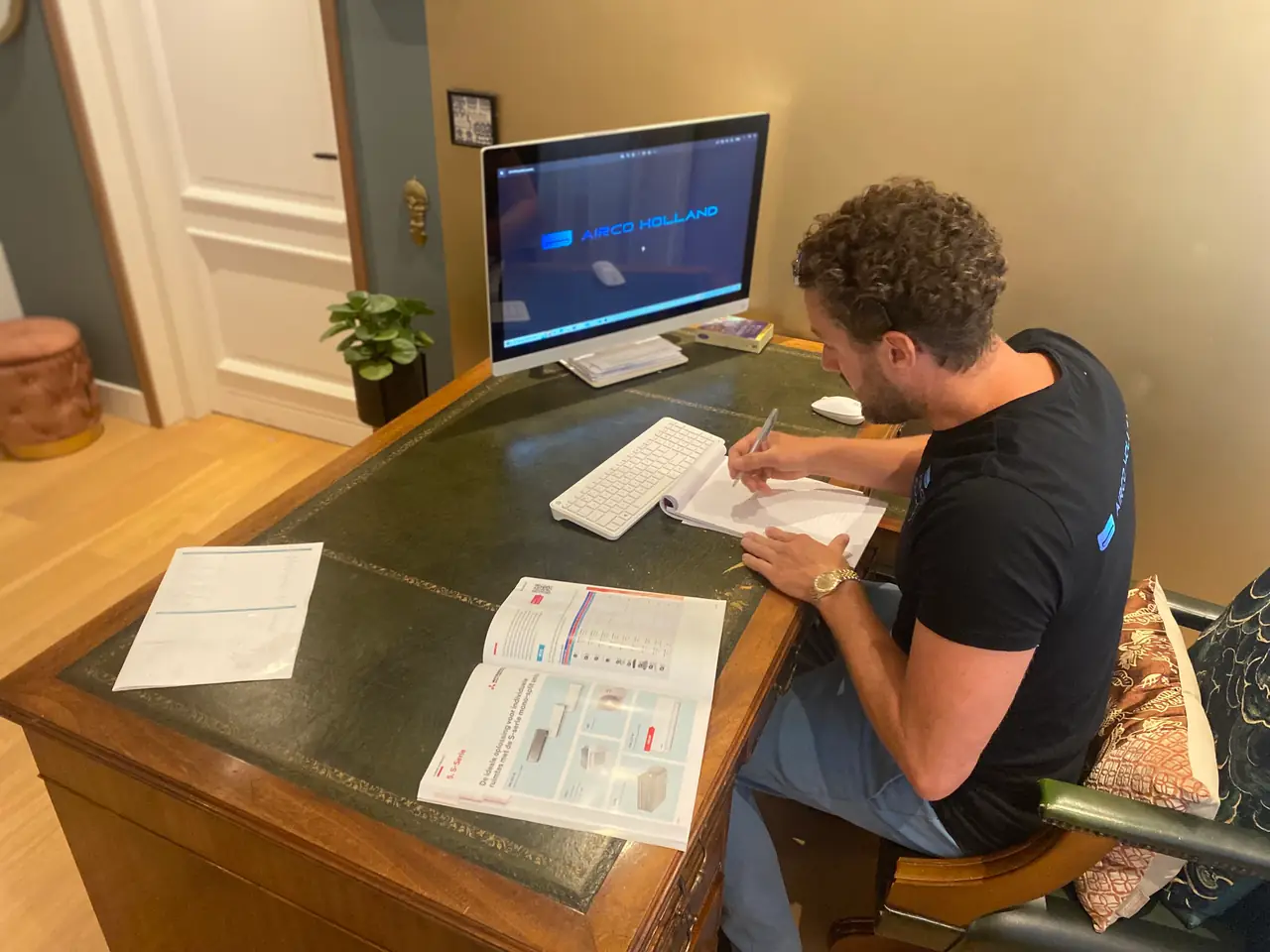 Rein schrijft aan een bureau met de brochure open, met Airco Holland op het scherm.