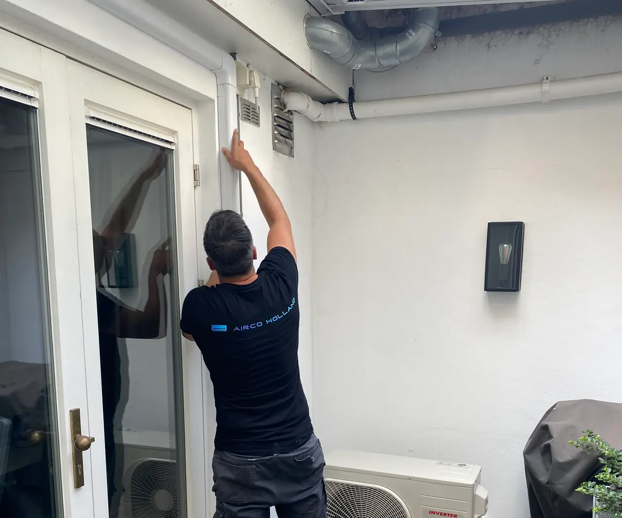 Peter werkt aan de leiding boven een tuindeur; een witte goot loopt strak langs muur en plafond.