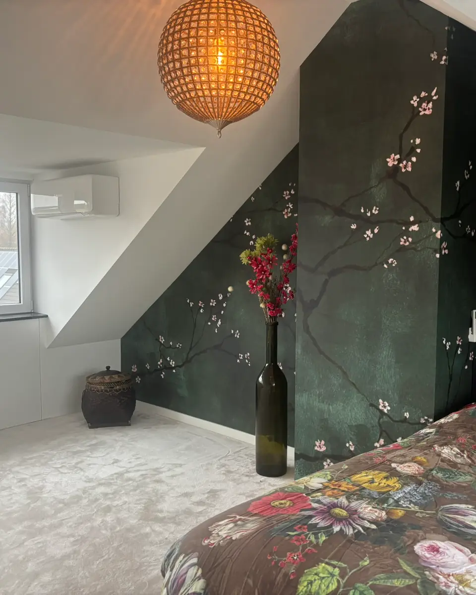 Een zolderslaapkamer met donkergroen bloesembehang, een rotan hanglamp en een wandmodel bij het raam.