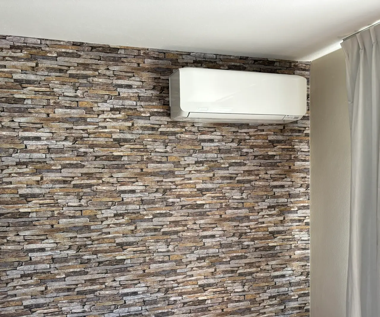 Een wit wandmodel hoog op een wand met steenstrips in een slaapkamer, naast een licht gordijn.