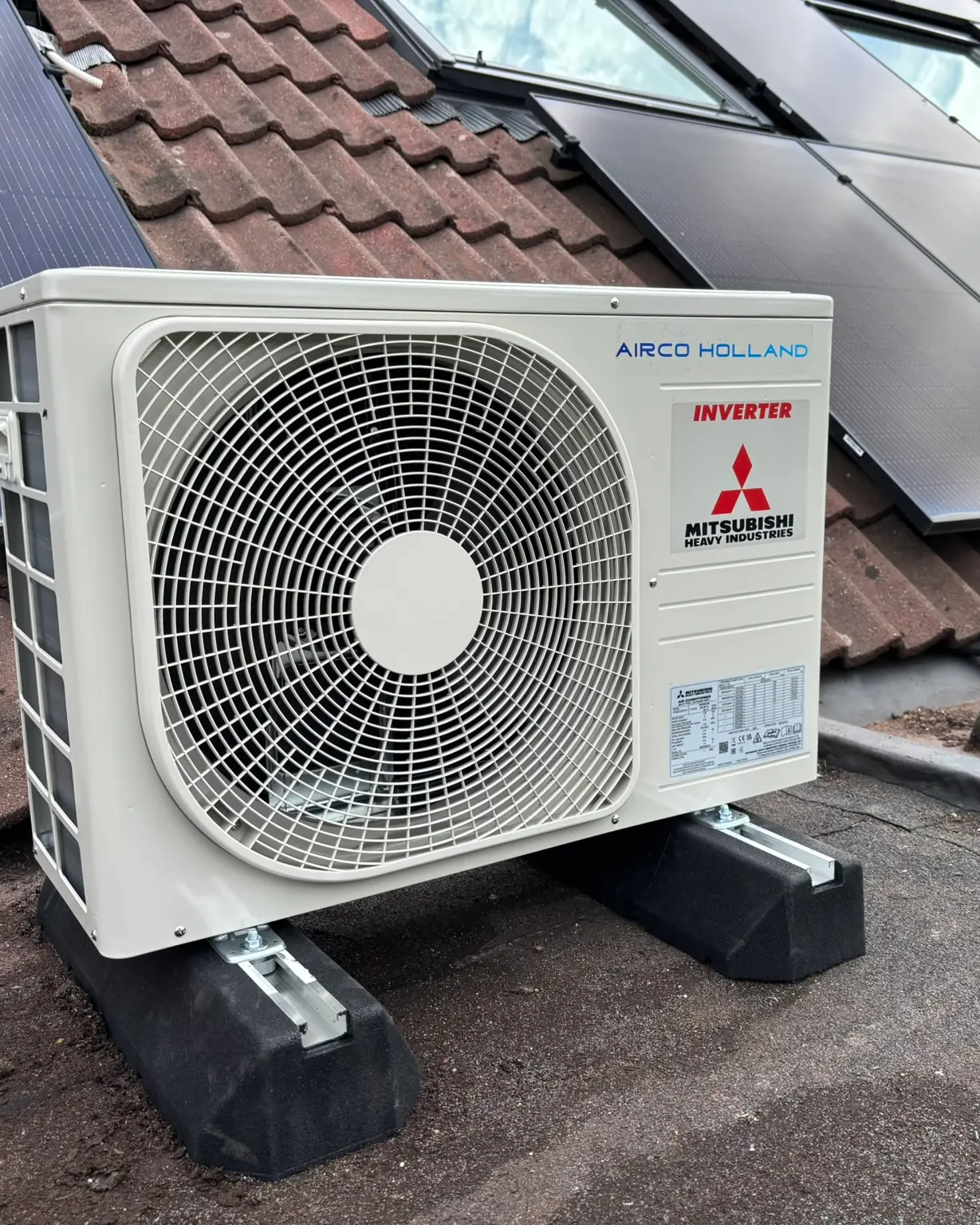 Een buitenunit op een plat dak naast een pannendak met zonnepanelen.