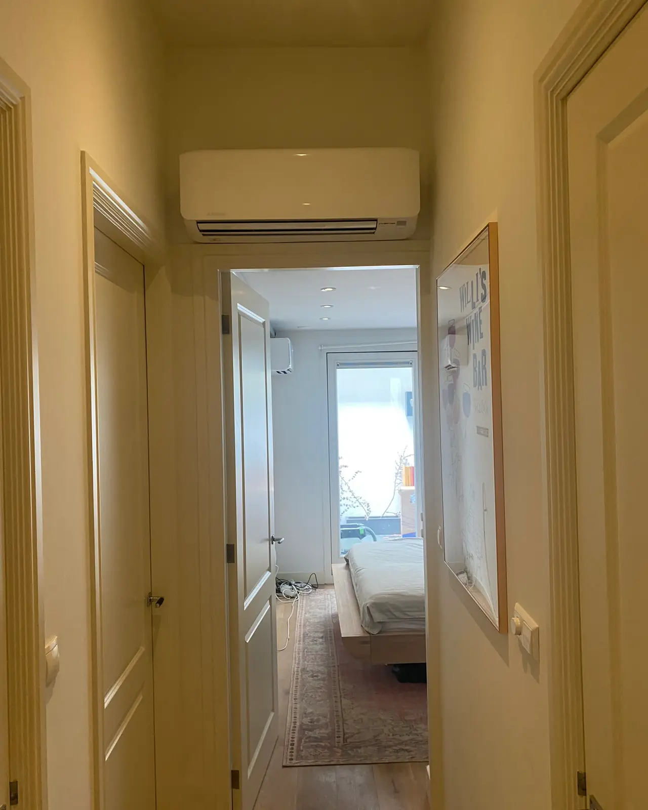 Een gang met een wit wandmodel boven de deur in warm lamplicht, met een doorkijk naar de slaapkamer.