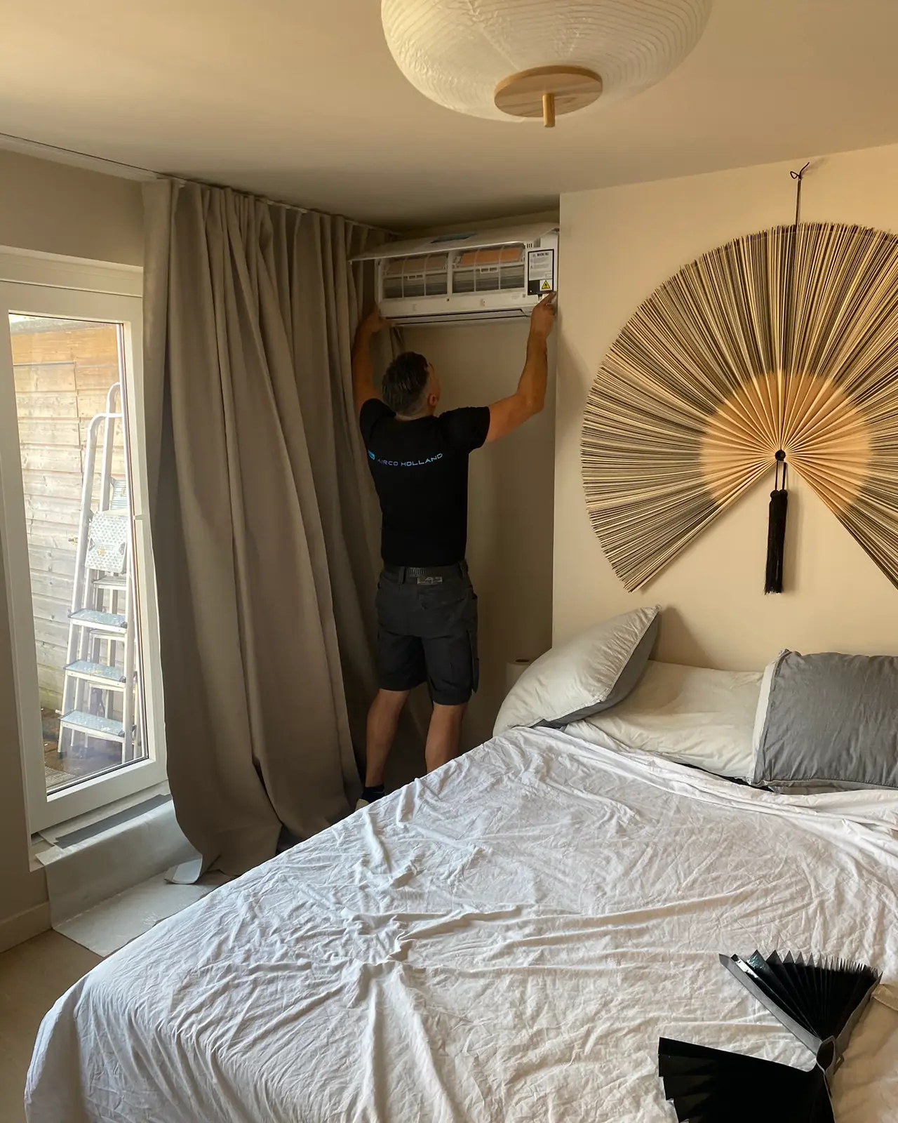 Peter hangt een wandmodel op in een slaapkamer met een grote waaier aan de muur; het bed is afgedekt.