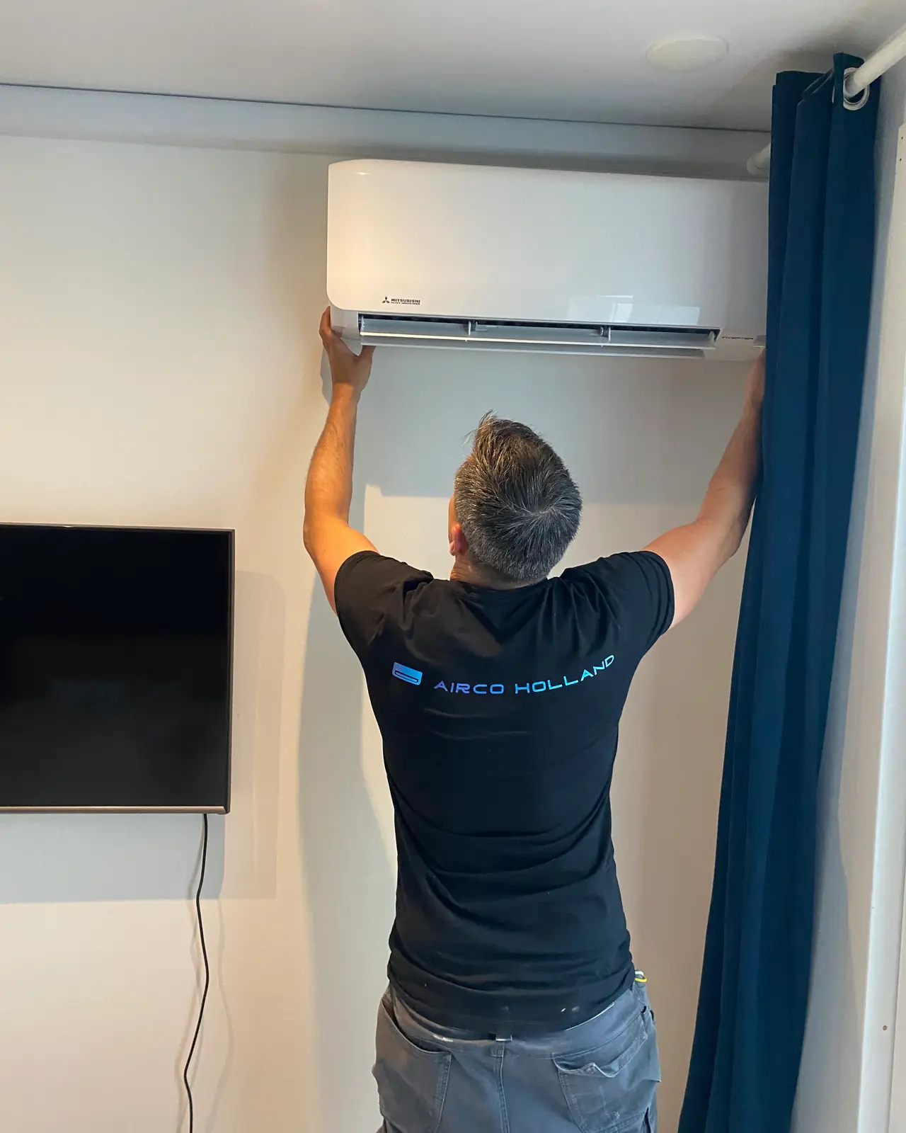 Peter hangt een wit wandmodel op naast een blauw gordijn en een tv.