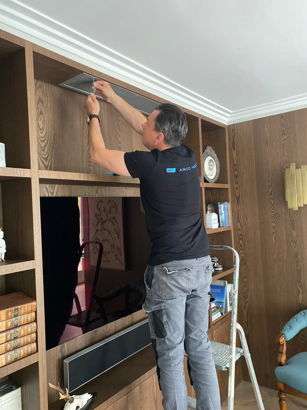 Peter of Airco Holland stands on a step and fits the air conditioner's grille at the top of a fitted cabinet.