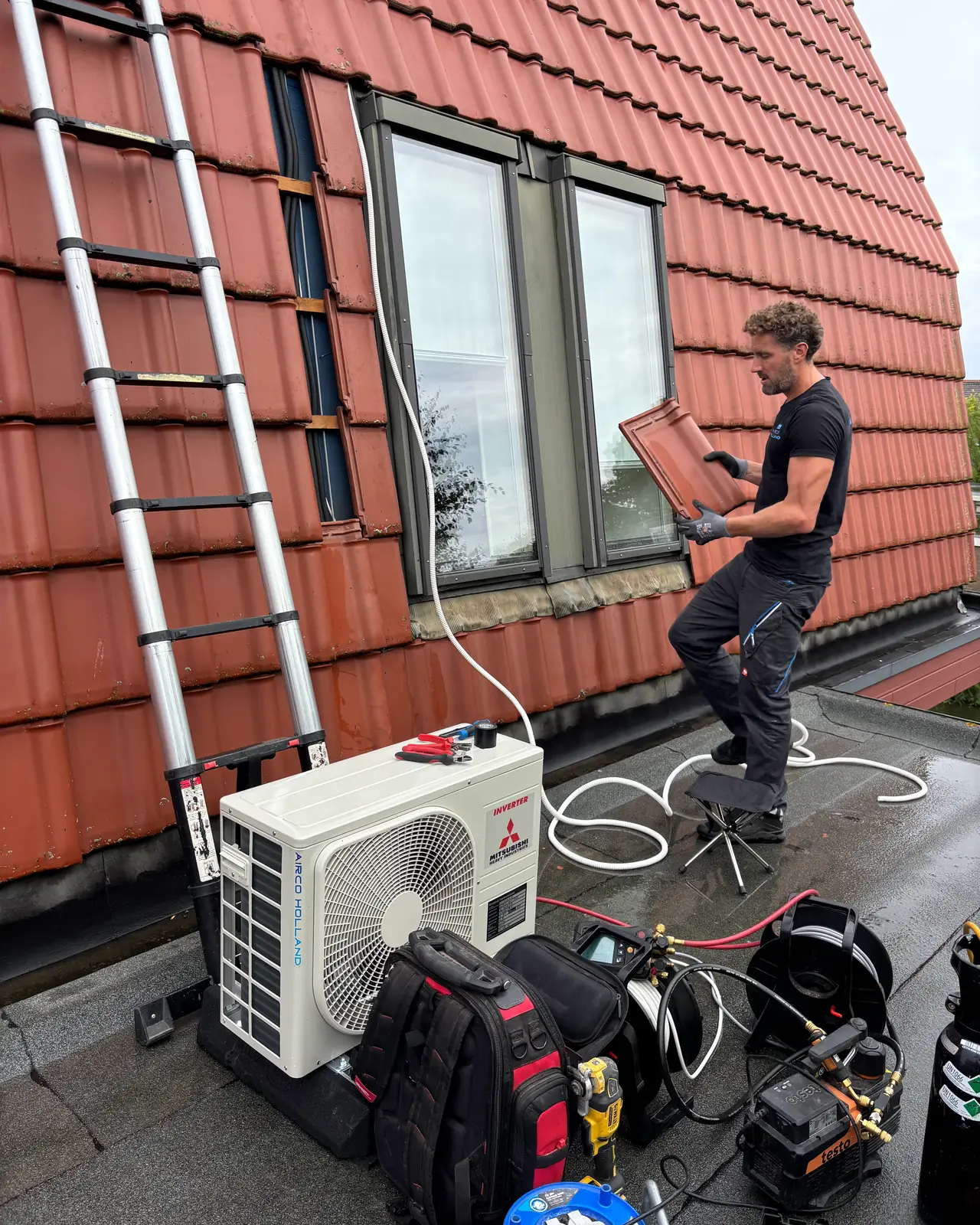 Rein houdt een losse dakpan vast bij een raam in een pannengevel; een witte leiding loopt naar het buitendeel.