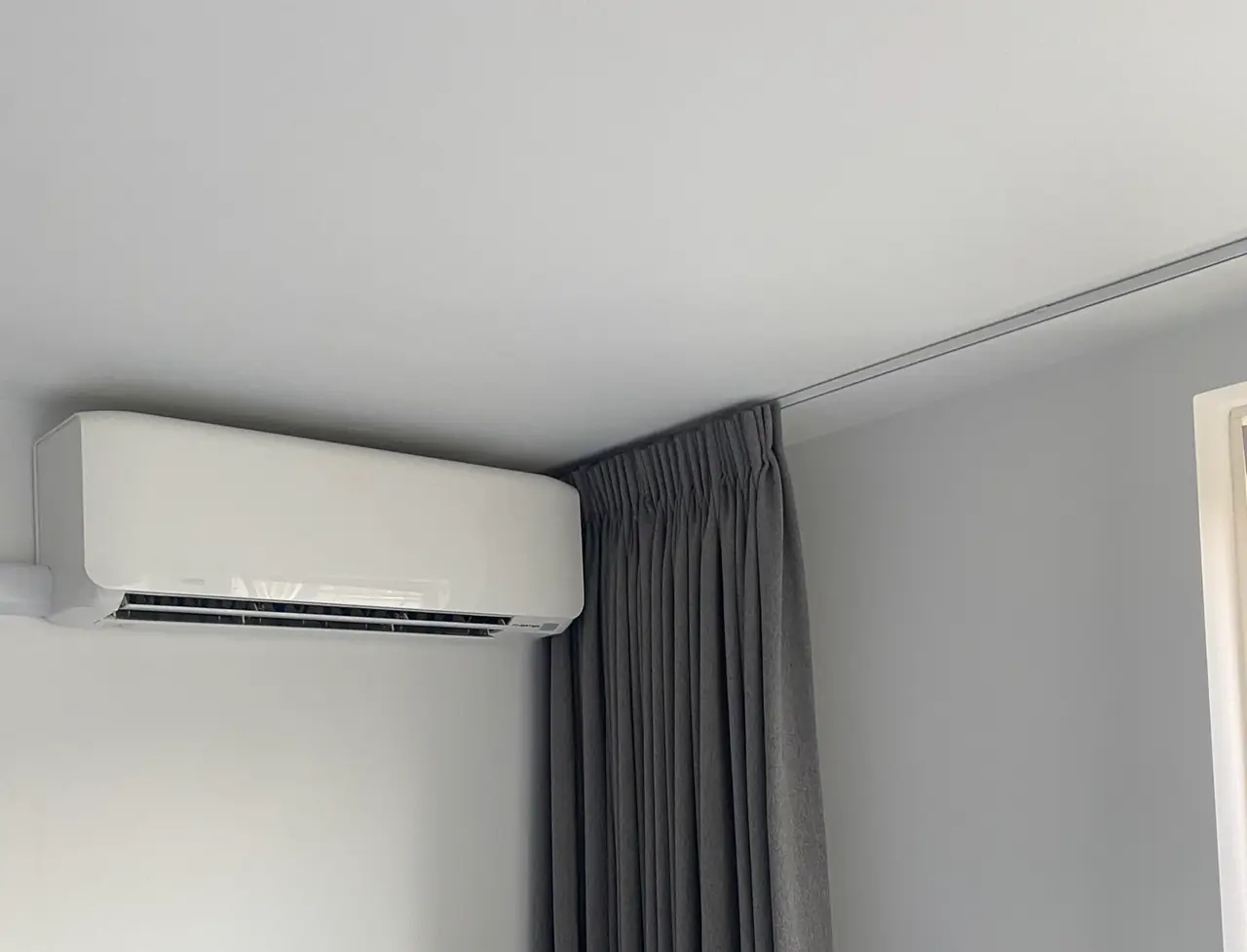Een wit wandmodel in de hoek van een kamer, strak onder het plafond naast een grijs gordijn.