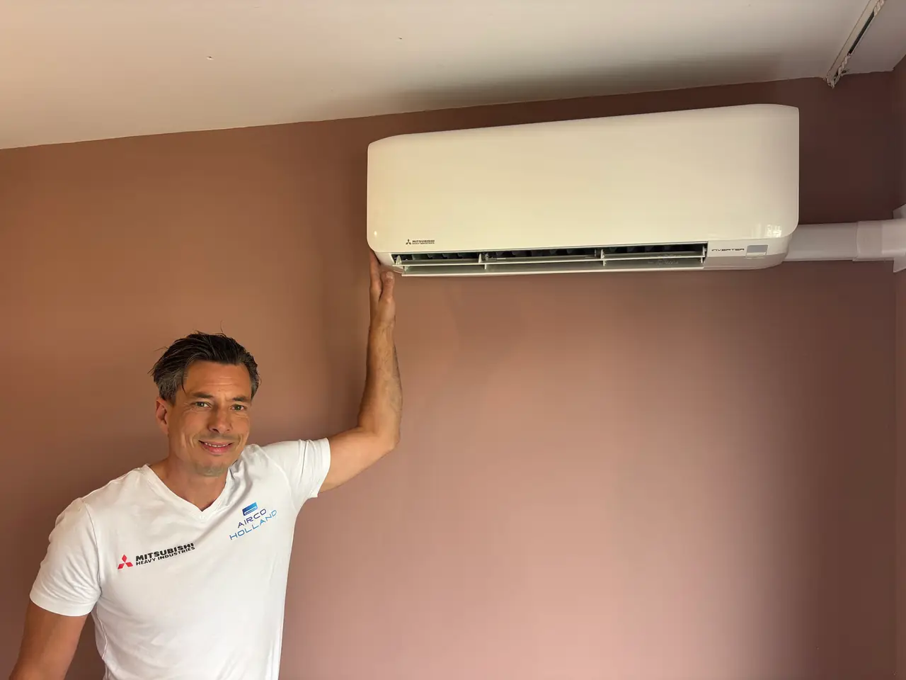 Peter, in een wit shirt, houdt zijn hand bij de uitblaas van een nieuw wit wandmodel aan een oudroze muur.