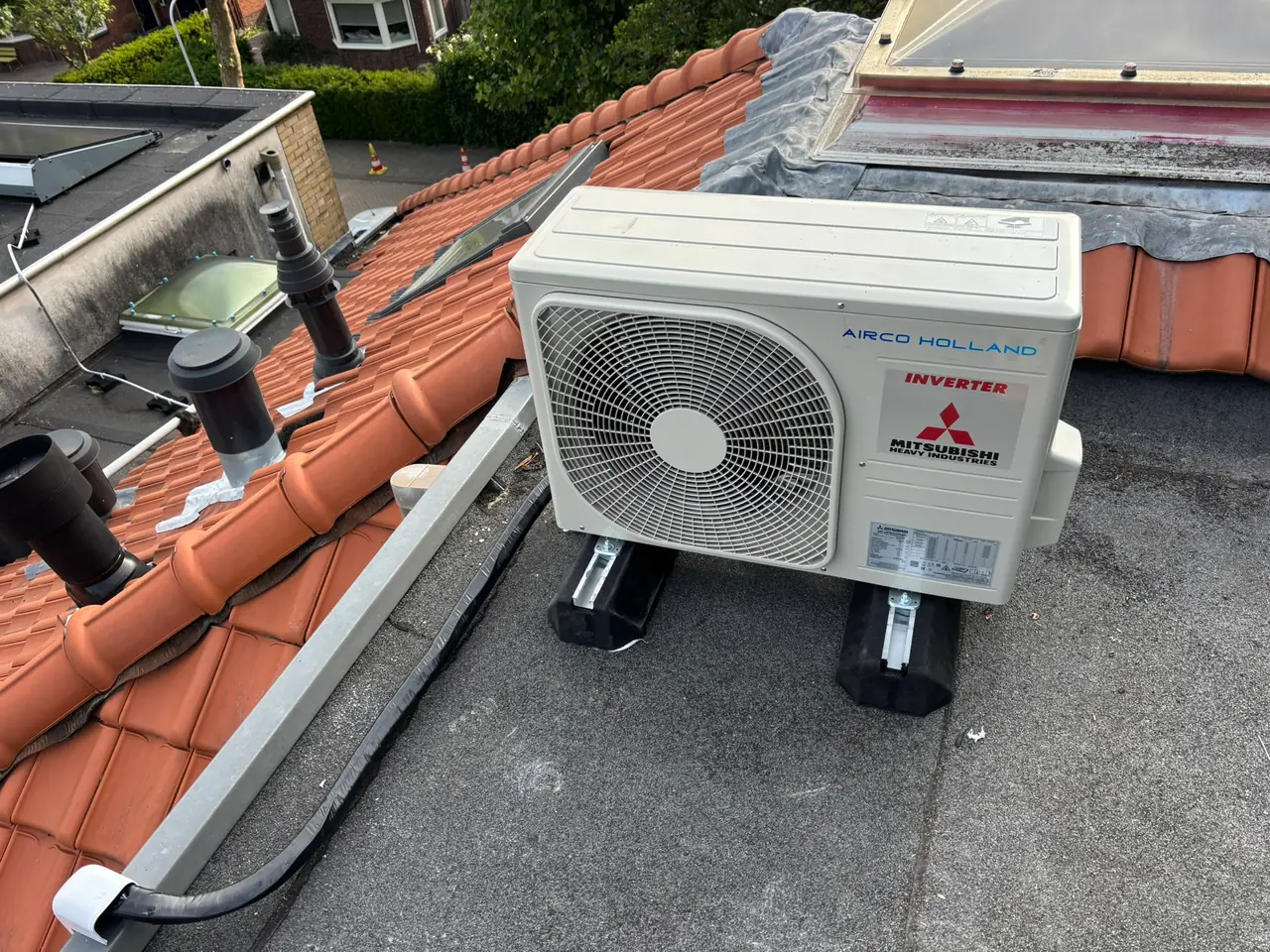 Een nieuwe buitenunit op dakvoeten op een plat dak, naast oranje dakpannen.
