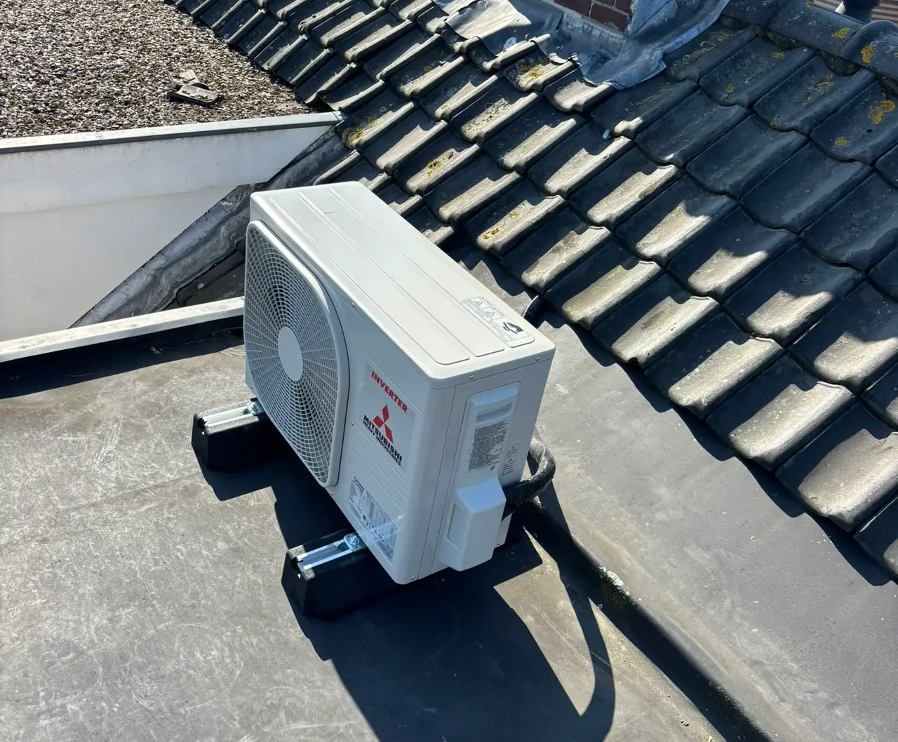 A new outdoor unit on roof feet in the sun, on a flat roof next to a dark tiled roof.