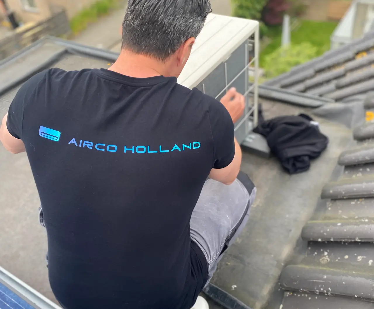 Peter, met Airco Holland op zijn rug, werkt aan een buitenunit op een plat dak.