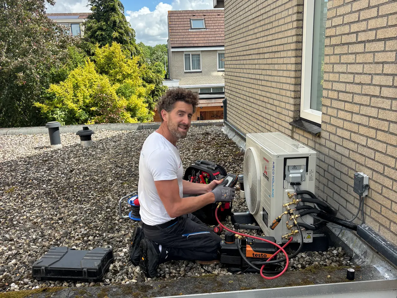 Rein knielt op een grinddak bij een nieuw buitendeel, met een meter in zijn hand en buurhuizen erachter.